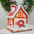 Decorative gingerbread house with red roof and lights, resembling a small house, on a white surface with a blurred green background.