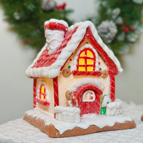 Decorative gingerbread house with red roof and lights, resembling a small house, on a white surface with a blurred green background.